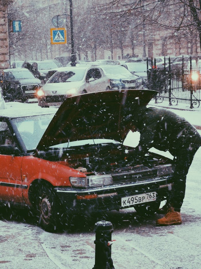 A person repairs a car engine on a snowy street in winter, showcasing auto maintenance challenges.