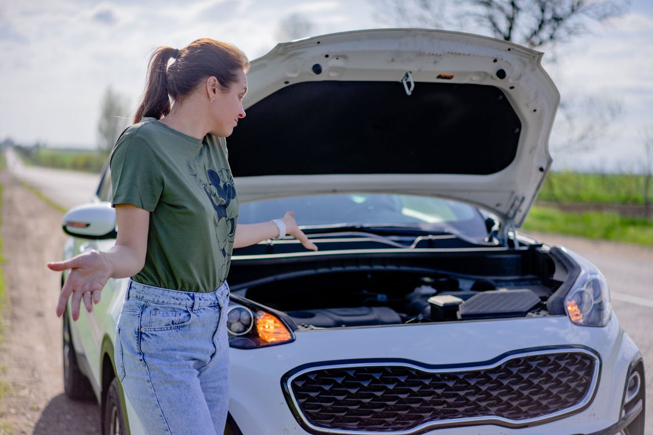 A woman looks puzzled by her broken car with an open hood on a rural road.